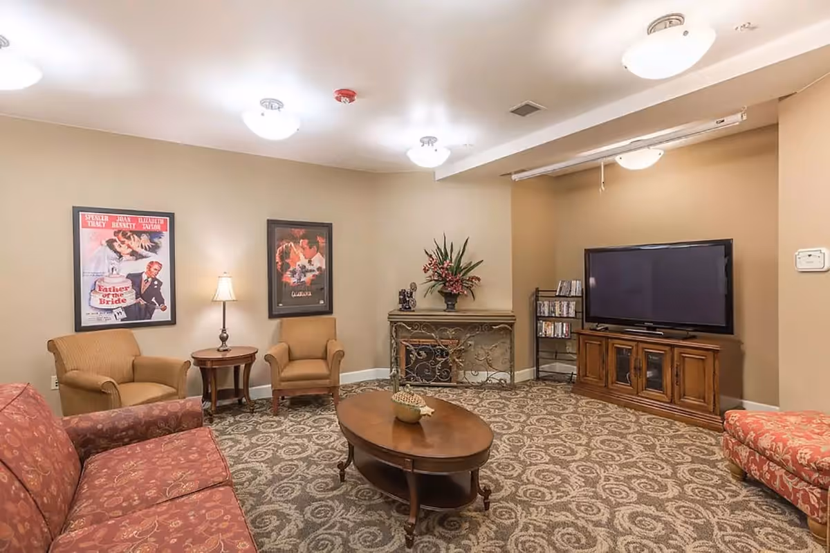A cozy living room with patterned carpet, two armchairs, a red floral sofa, a wooden coffee table with a decorative bowl, a TV on a wooden stand, and framed movie posters on the beige walls. There is also a decorative console table with a flower arrangement and a rack with DVDs.