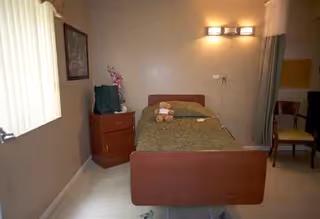 A bedroom in a senior care facility featuring a single bed, dresser, and window blinds.