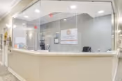 Reception desk area with a large glass partition, a computer monitor, office chair, and a sign on the wall behind the desk in a well-lit interior space.
