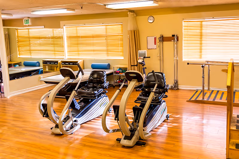 A rehabilitation exercise room with two recumbent exercise bikes in the center, wooden flooring, large windows with blinds, a therapy table with a blue bolster, a mirror on one wall, and parallel bars for walking exercises.