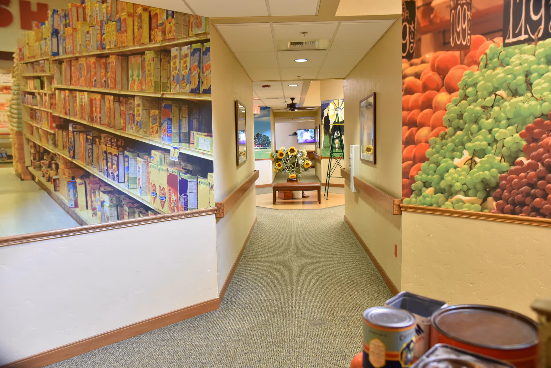 A hallway in a senior living facility with large wall murals depicting grocery store shelves stocked with cereal boxes on the left and fresh fruits including grapes on the right. At the end of the hallway, there is a small table with a vase of sunflowers and a television mounted on the wall.