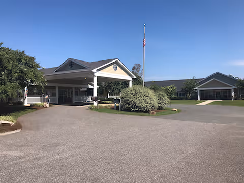 Front entrance of a senior living facility with a covered porte-cochere, flagpole, and driveway under a clear blue sky.