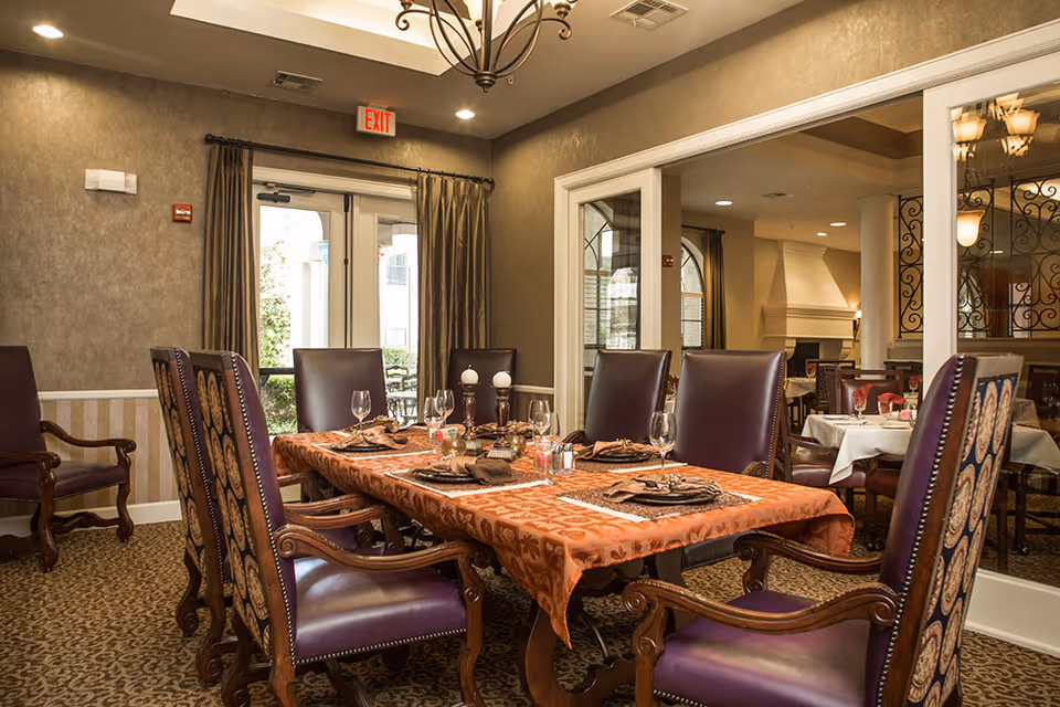 Dining room with an orange tablecloth-covered table set with glassware and surrounded by upholstered wooden chairs under a chandelier.