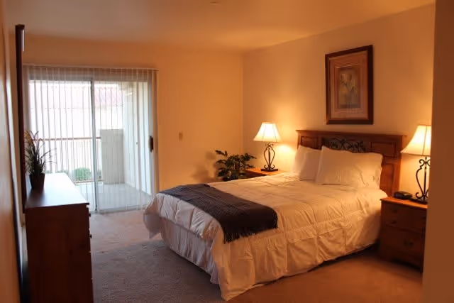 A cozy bedroom with a neatly made bed featuring white bedding and a dark throw blanket. There are two bedside tables with lamps on each side of the bed, a framed picture on the wall above the headboard, a wooden dresser with a plant on top, and sliding glass doors leading to an outdoor balcony or patio.