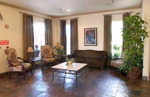 Cozy senior living room with seating area, coffee table, and large plants by windows.