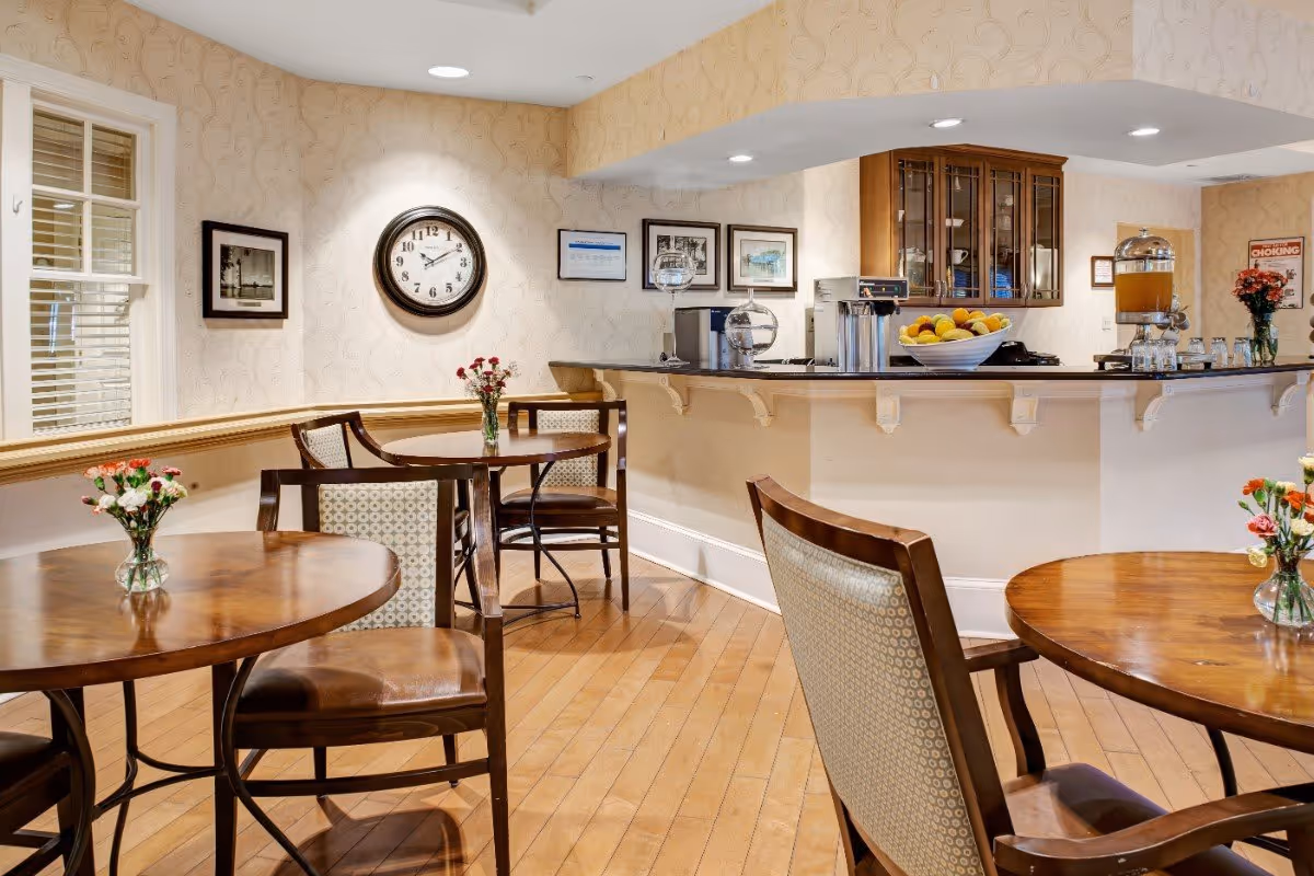 Bright dining/lounge area with round wooden tables and chairs, a service counter with fruit bowl and beverage dispensers, and a wall clock and framed pictures.