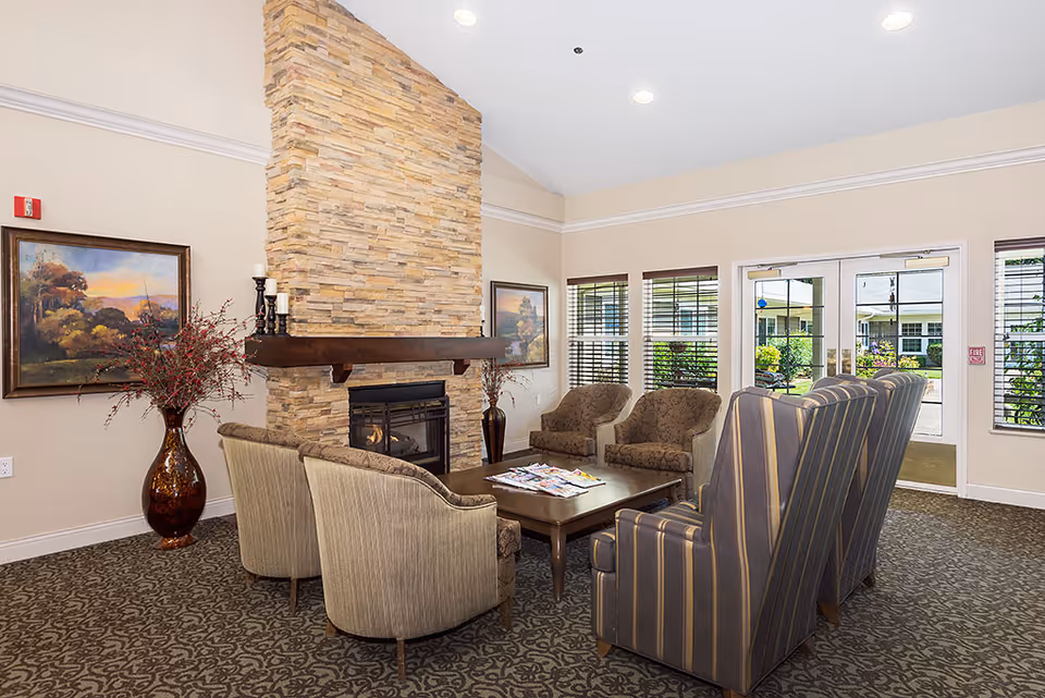 Comfortable seating arranged around a stone fireplace in a bright retirement community lounge with large windows and double doors.