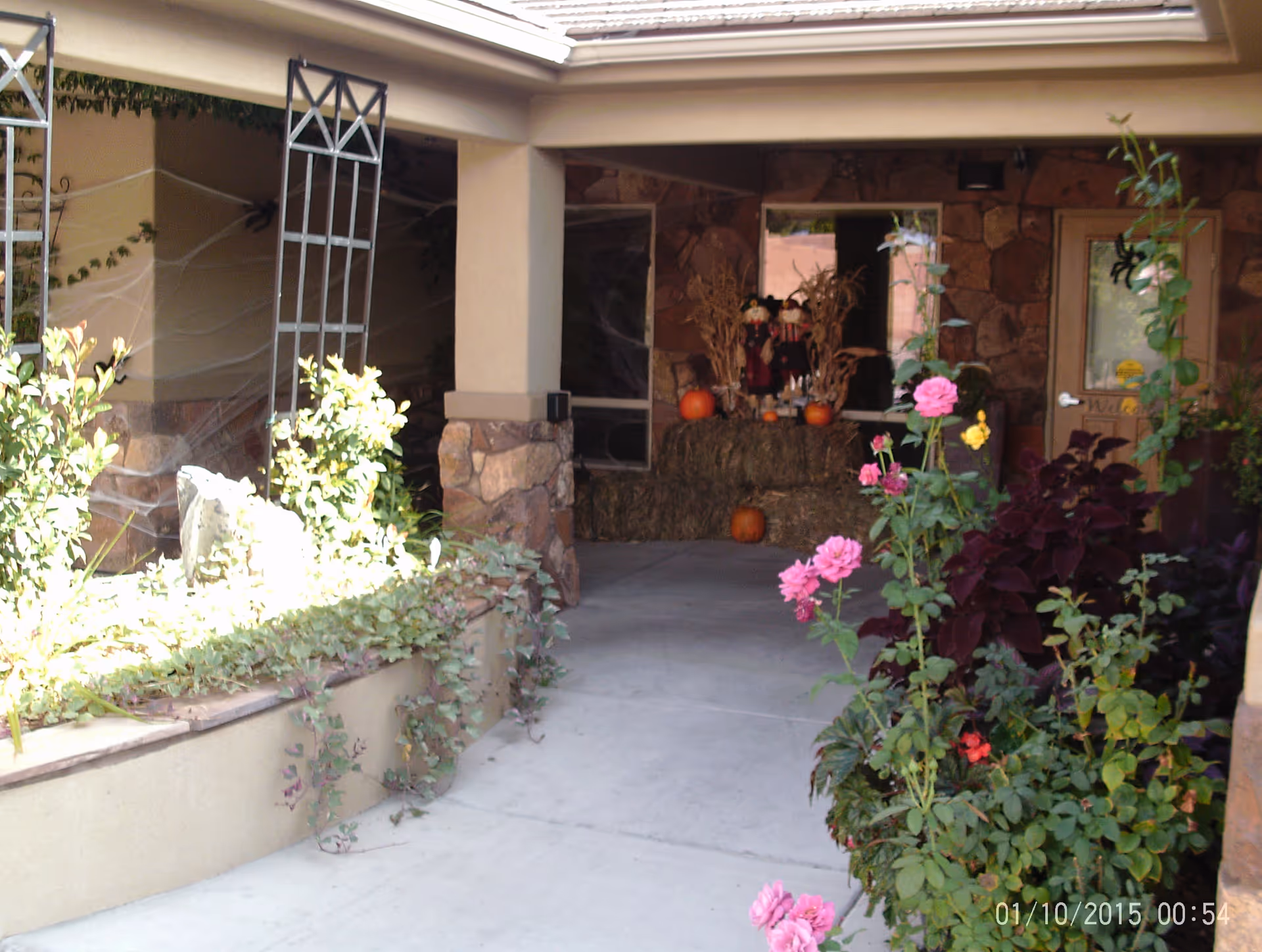 Covered outdoor walkway with stone pillars and a garden bed filled with green plants and pink flowers. At the end of the walkway, there are hay bales decorated with pumpkins and scarecrow figures, suggesting a fall or Halloween theme.