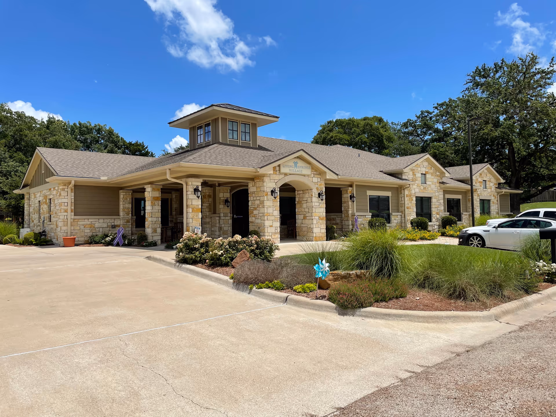 Stone-faced single-story building with an arched covered entrance, landscaped driveway and parked cars under a blue sky.