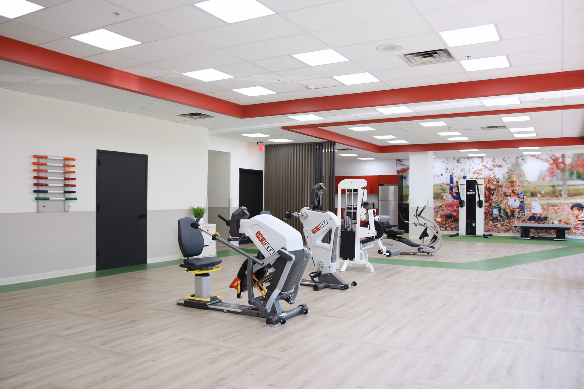 A spacious rehabilitation gym area with exercise equipment including seated machines and a recumbent bike. The room has light wood flooring, white walls with a red ceiling border, and a large mural of children playing in leaves on one wall. There are black doors and a rack with colored rods on one wall.