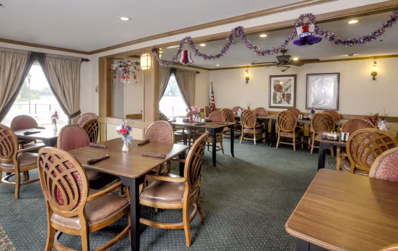 A dining room with several wooden tables and chairs arranged neatly. Each table has a small floral centerpiece and rolled napkins. The room is decorated with garlands hanging from the ceiling and has large windows with curtains allowing natural light to enter. There are framed pictures on the walls and an American flag in the corner.