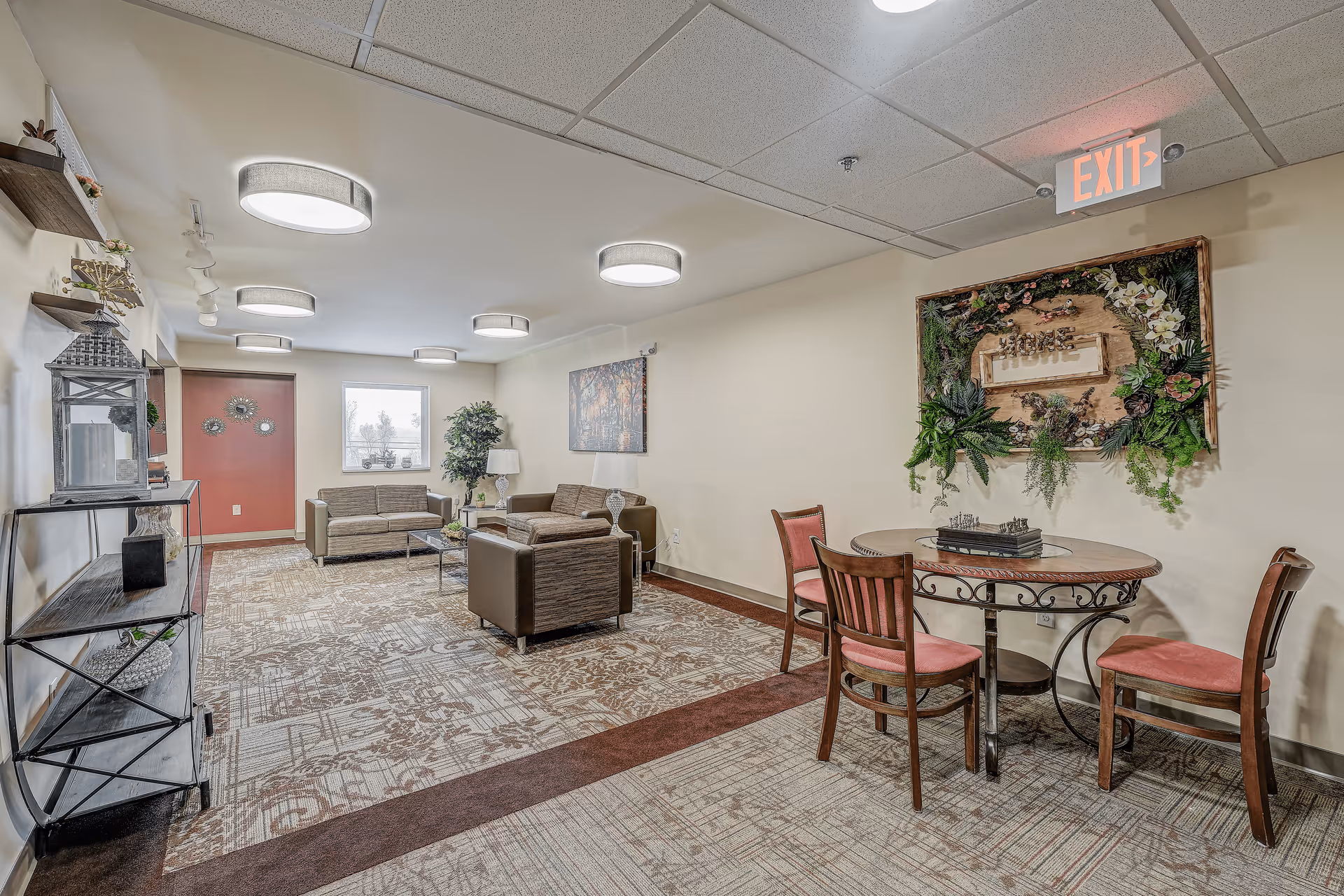 A cozy common area in a senior living facility featuring two sofas and an armchair arranged around a glass coffee table on a patterned carpet. To the right, there is a round table with three wooden chairs with red cushions. The walls are decorated with a large framed floral artwork and a painting. Shelves with decorative items are mounted on the left wall. Ceiling lights illuminate the space, and an exit sign is visible above the floral artwork.