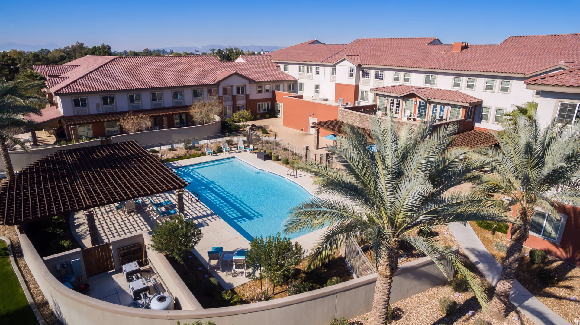Aerial view of an outdoor swimming pool area at a senior living facility with lounge chairs around the pool, palm trees, and a pergola providing shade. Surrounding the pool are multi-story buildings with red tile roofs and beige and red exterior walls under a clear blue sky.