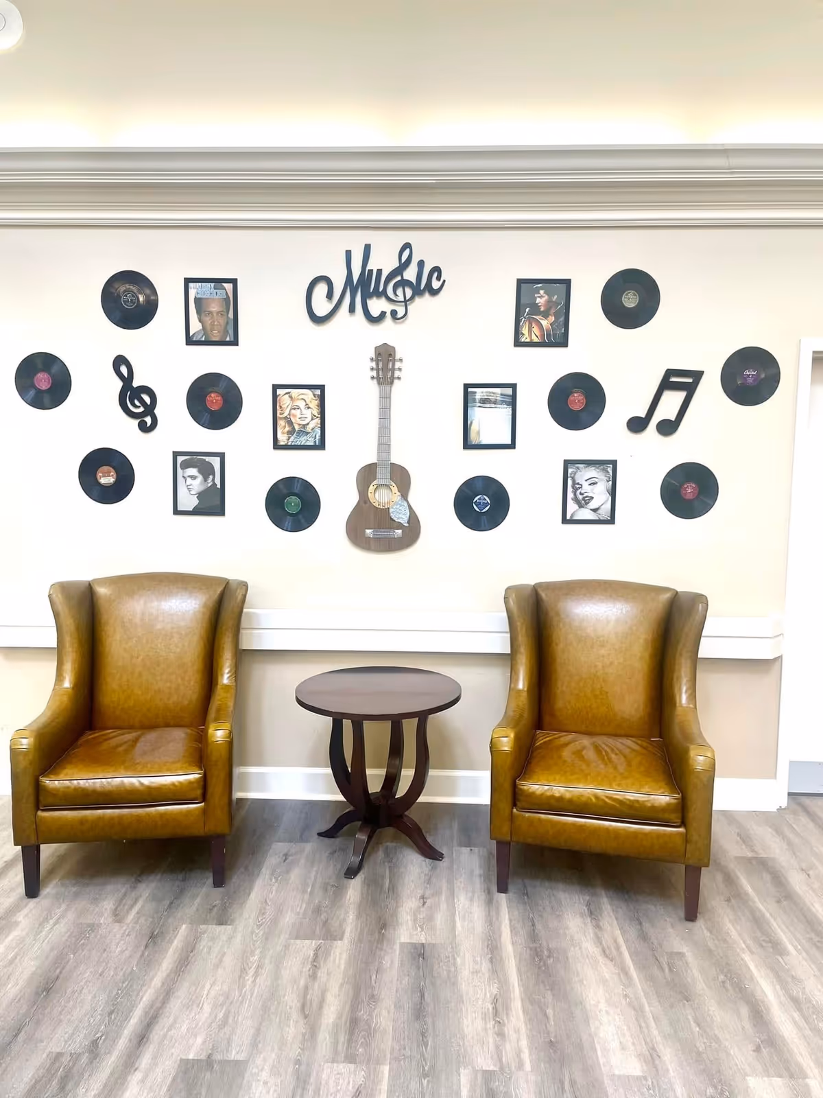 Two brown leather armchairs with a small round wooden table between them, set against a wall decorated with vinyl records, framed pictures of famous musicians, a wooden guitar, and musical note decorations with the word 'Music' displayed prominently.