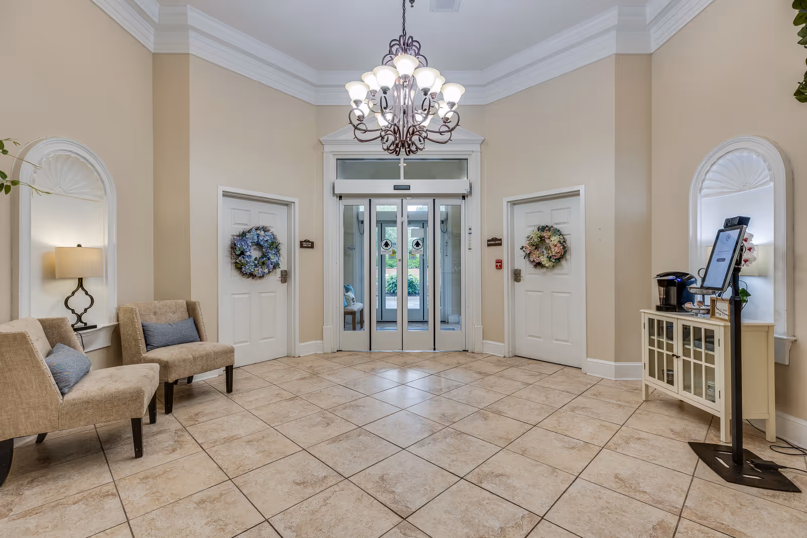 A bright and spacious entrance lobby with beige tiled floors and cream-colored walls. Two beige upholstered chairs with blue cushions are placed on the left side, next to a wall lamp. Two white doors adorned with floral wreaths flank a glass double door leading outside. A chandelier with multiple lights hangs from the ceiling. On the right side, there is a small white cabinet with a coffee maker and a digital sign stand.