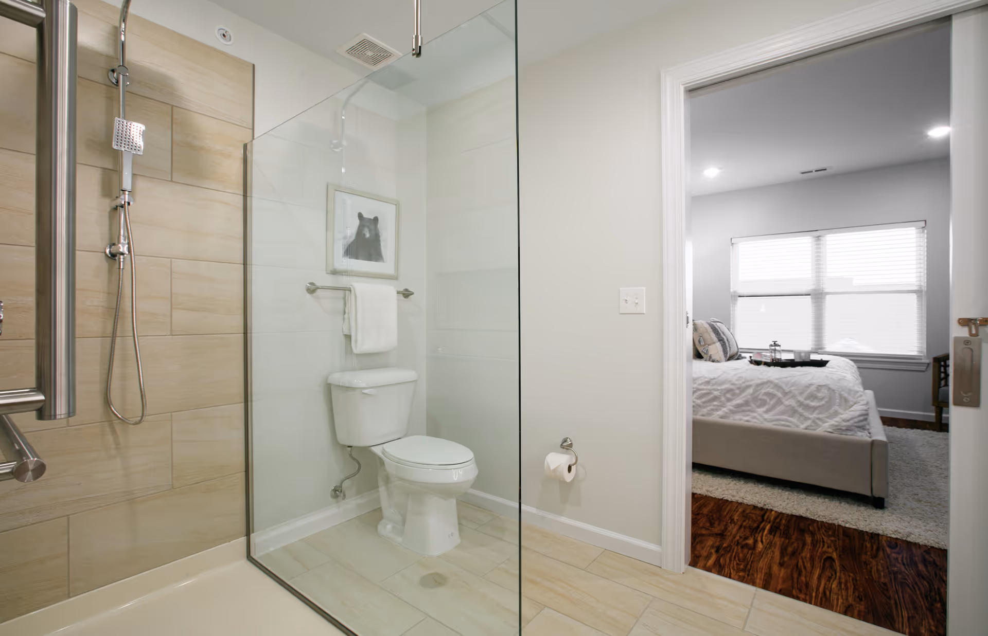 Modern bathroom with a glass-enclosed shower, toilet, towel rack and a view into an adjacent bedroom.