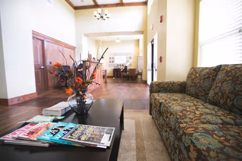 A cozy interior common area with a patterned sofa on the right, a black coffee table with magazines and a vase of decorative flowers, wooden floors, and a dining area visible in the background under warm lighting.