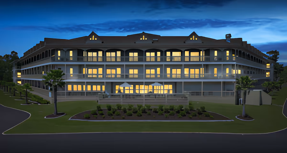 Exterior view of a large multi-story building at dusk with many windows illuminated from inside. The building is surrounded by a well-maintained lawn, small trees, and landscaped garden beds. The sky is dark blue with some clouds.