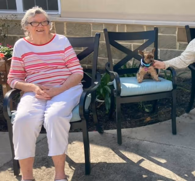 An elderly woman wearing glasses and a striped pink and white shirt sits on a cushioned outdoor chair next to a small dog sitting on another cushioned chair. A person’s hand is reaching out to pet the dog. The setting appears to be an outdoor patio area with a stone wall and plants in the background.