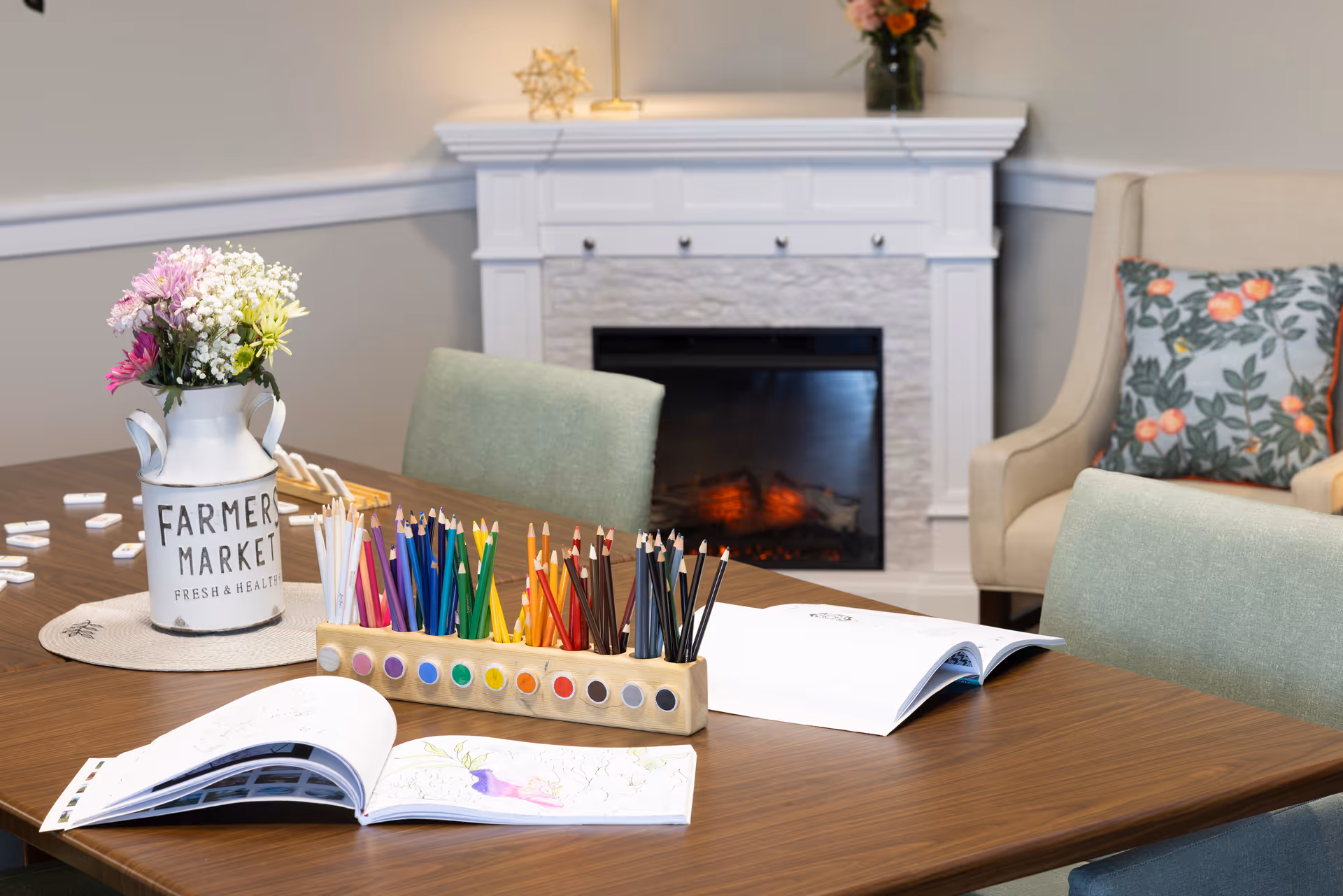 A cozy activity table with colored pencils, watercolor paints, and open coloring books in front of a fireplace and upholstered chairs.