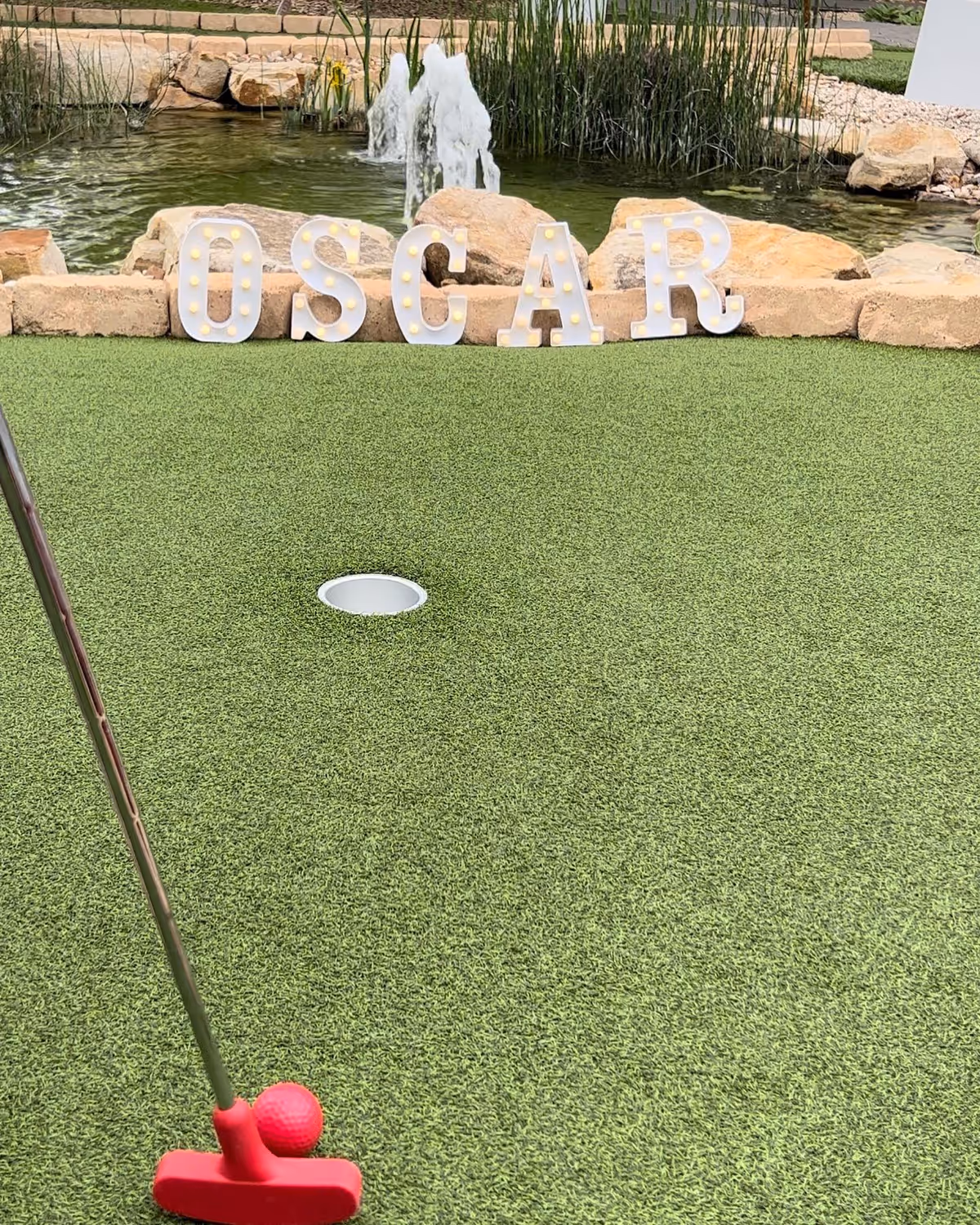 A mini golf putting green with a red putter and red golf ball near the hole. In the background, there is a small pond with a water fountain and rocks. Large white marquee letters spelling 'OSCAR' with lights are placed on the rocks by the pond.