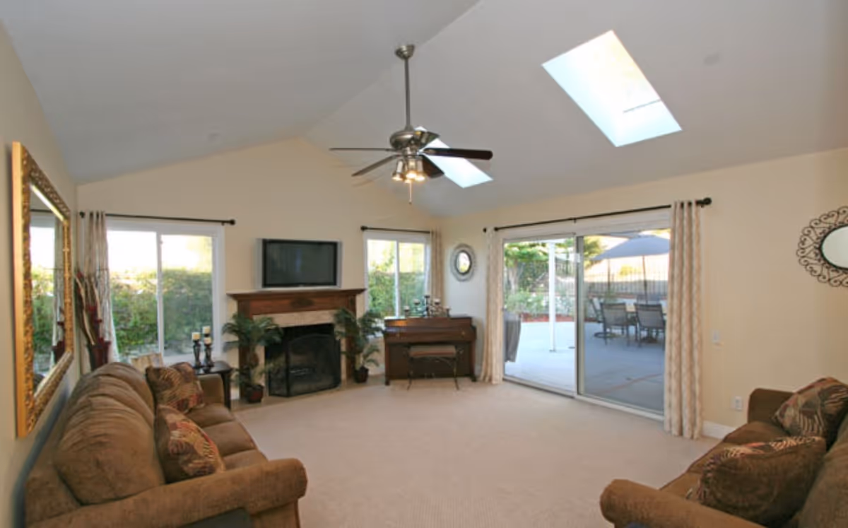 Spacious living room with vaulted ceiling, ceiling fan and skylights, sofas, a fireplace and TV, and sliding glass doors to an outdoor patio.