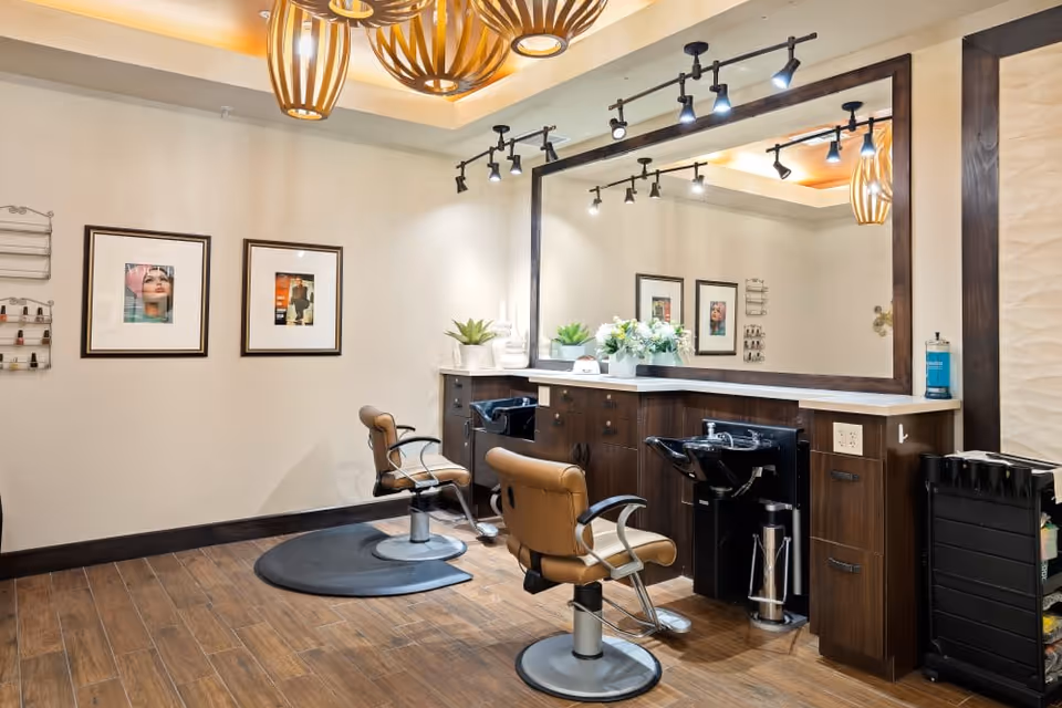 Interior view of a salon area with two brown salon chairs in front of a large mirror. The space features wooden flooring, dark wood cabinetry with sinks, decorative plants, framed pictures on the wall, and modern pendant lighting hanging from the ceiling.