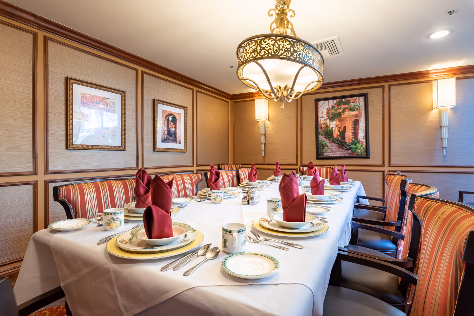 A formal dining room with a long table set for a meal, red folded napkins, china place settings, and striped chairs under a chandelier.