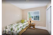 A small bedroom with beige walls and a single bed covered with a green and white floral bedspread. Next to the bed is a wooden nightstand with a lamp and a small plant. A window with white blinds lets in natural light, and there is a white closet door on the right side of the room.