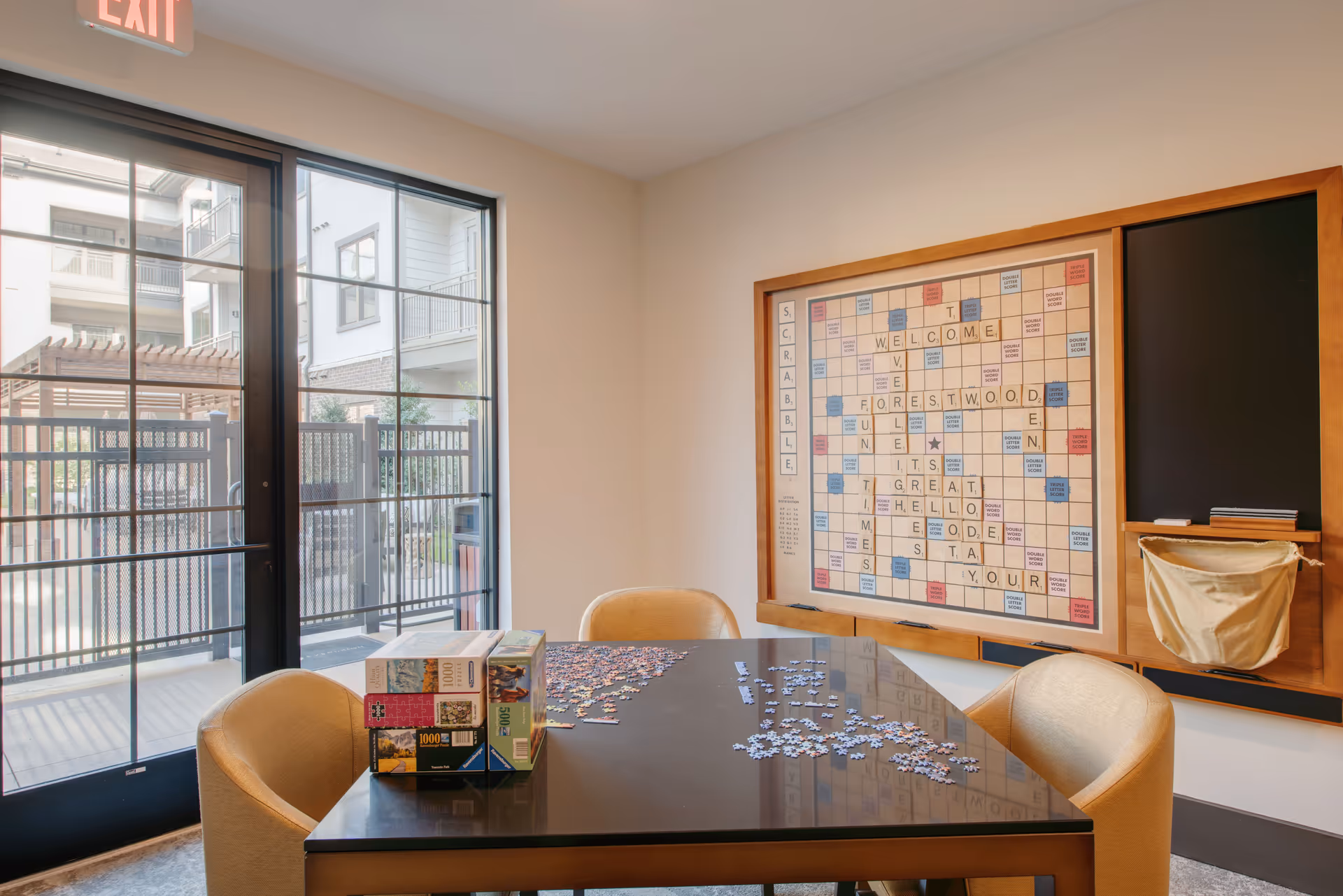 A small game room with a table covered in puzzle pieces, chairs, and a large wall-mounted Scrabble board beside a glass exit door.