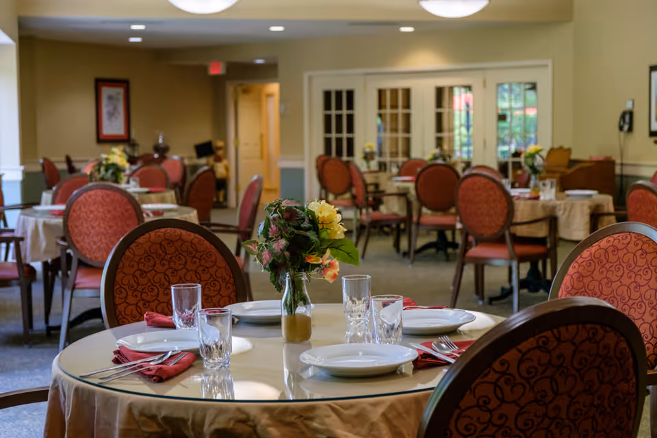 A dining room in a senior living facility with round tables covered in beige tablecloths. Each table is set with white plates, glasses, silverware, and red napkins. There are floral centerpieces on the tables. The room has red cushioned chairs with patterned backs, beige walls, and glass-paneled double doors in the background.