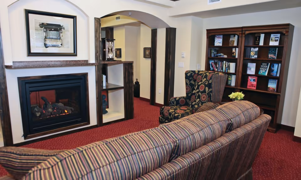 A cozy living room area with a striped sofa facing a fireplace with a framed picture of a typewriter above it. There is a patterned armchair next to a wooden bookshelf filled with books. The room has red carpet flooring and cream-colored walls with dark wooden trim.