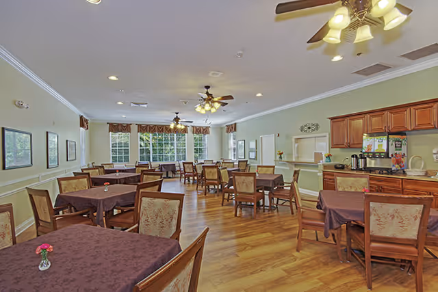 A spacious dining room with multiple tables covered in dark tablecloths, each surrounded by wooden chairs with cushioned seats and backs. The room has large windows letting in natural light, ceiling fans with lights, and a wooden floor. There is a counter area with coffee dispensers and cabinets along one wall.