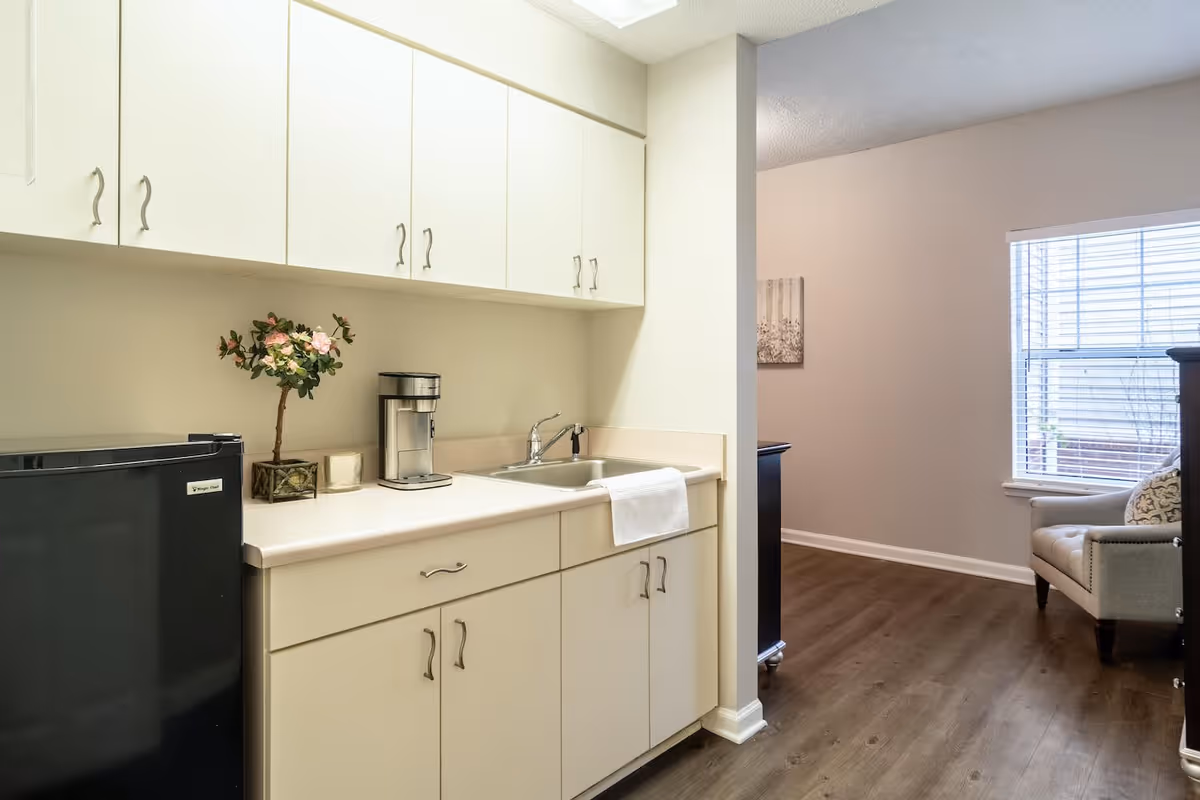 A small kitchenette area with white cabinets, a countertop with a coffee maker, a small potted plant, and a sink with a towel hanging on the edge. To the left is a black mini refrigerator. The kitchenette opens into a room with a window, a cushioned armchair, and wooden flooring.