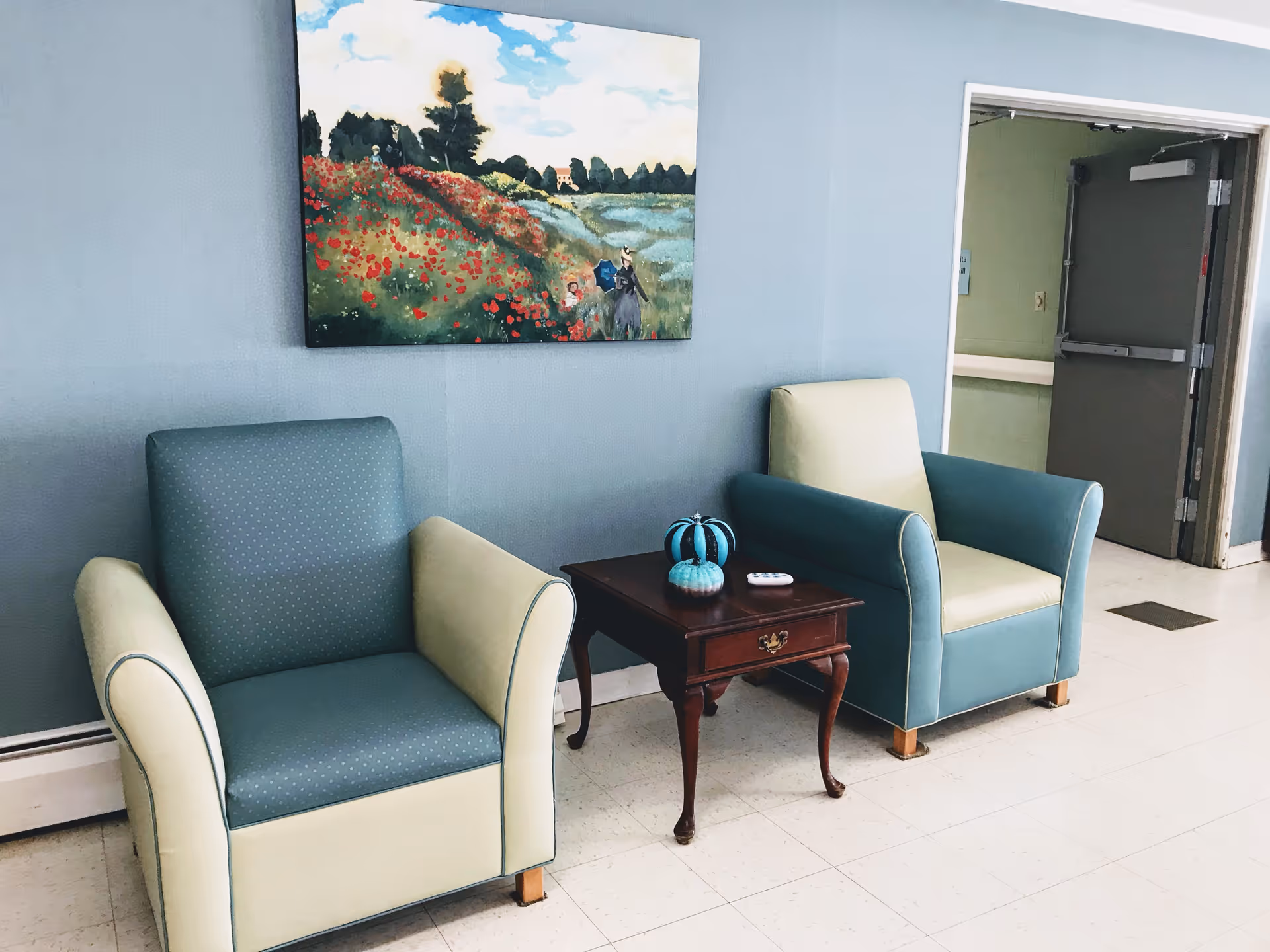 Two cushioned armchairs with blue and cream upholstery are placed on either side of a small wooden side table with decorative pumpkins and a remote control on top. The chairs and table are set against a light blue wall with a colorful painting of a landscape featuring a field of red flowers, trees, and a blue sky. To the right, there is an open doorway leading to a hallway with a gray door.