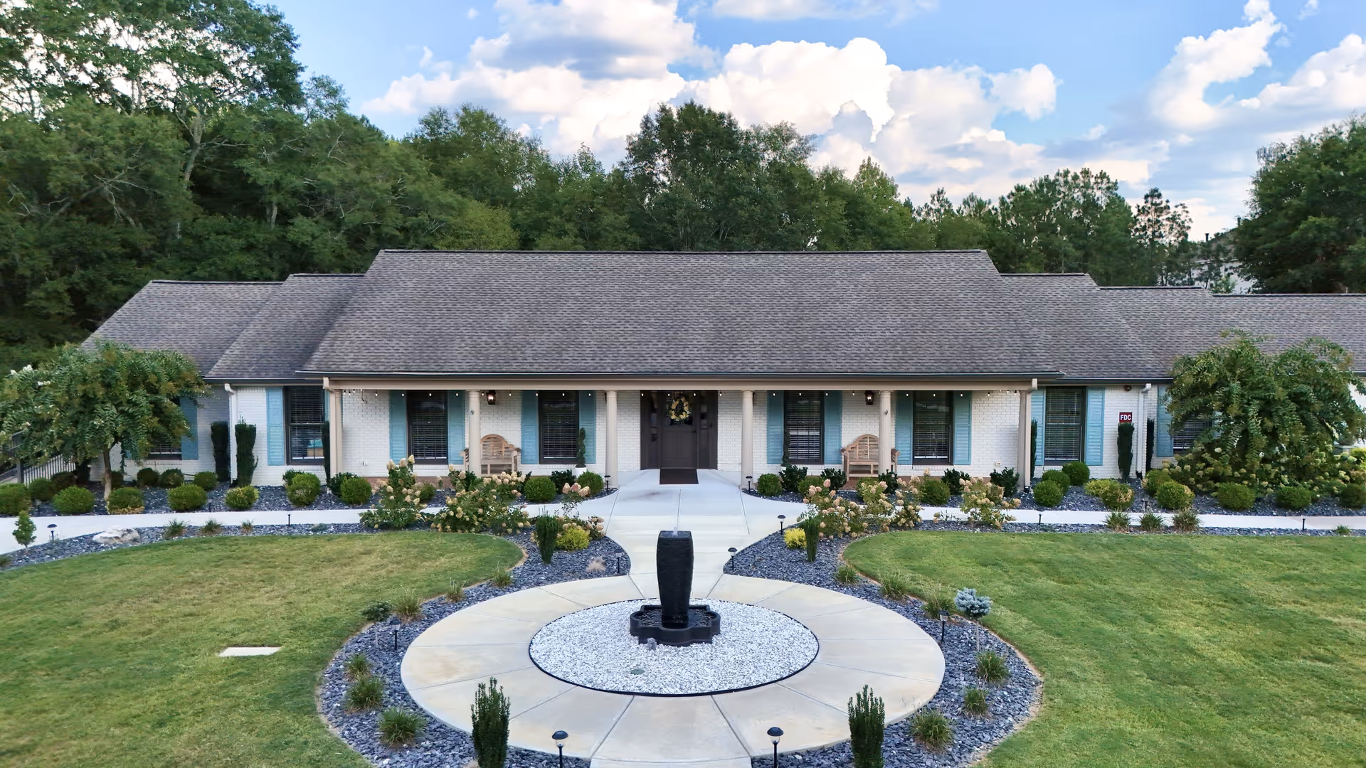 Single-story senior living building front with a central entrance, circular fountain, and manicured lawns and landscaping.