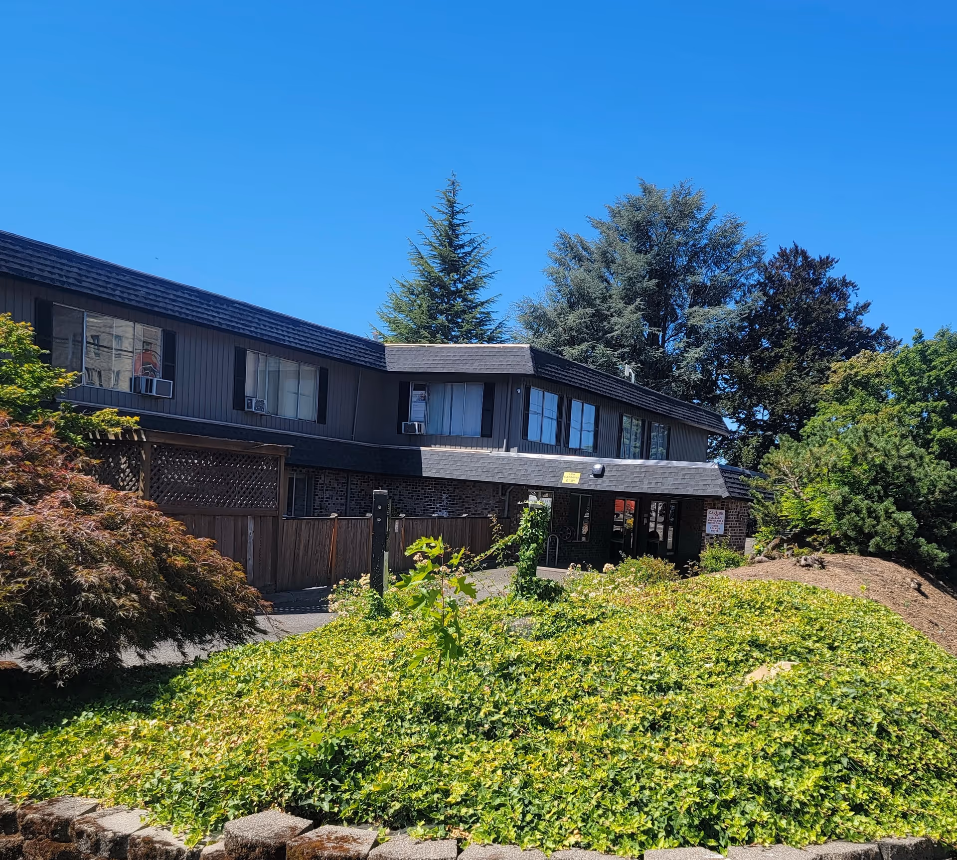 Two-story building front with an entrance, surrounding shrubs and trees, and a clear blue sky.