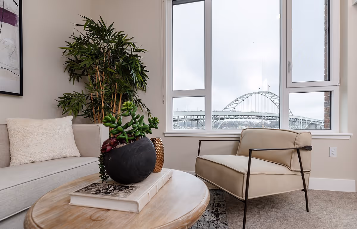 A cozy living room area with a beige sofa and a matching armchair with metal arms. A round wooden coffee table holds a large black planter with green succulents and a book. A tall green plant is placed in the corner near a large window, which offers a view of a bridge outside on a cloudy day.