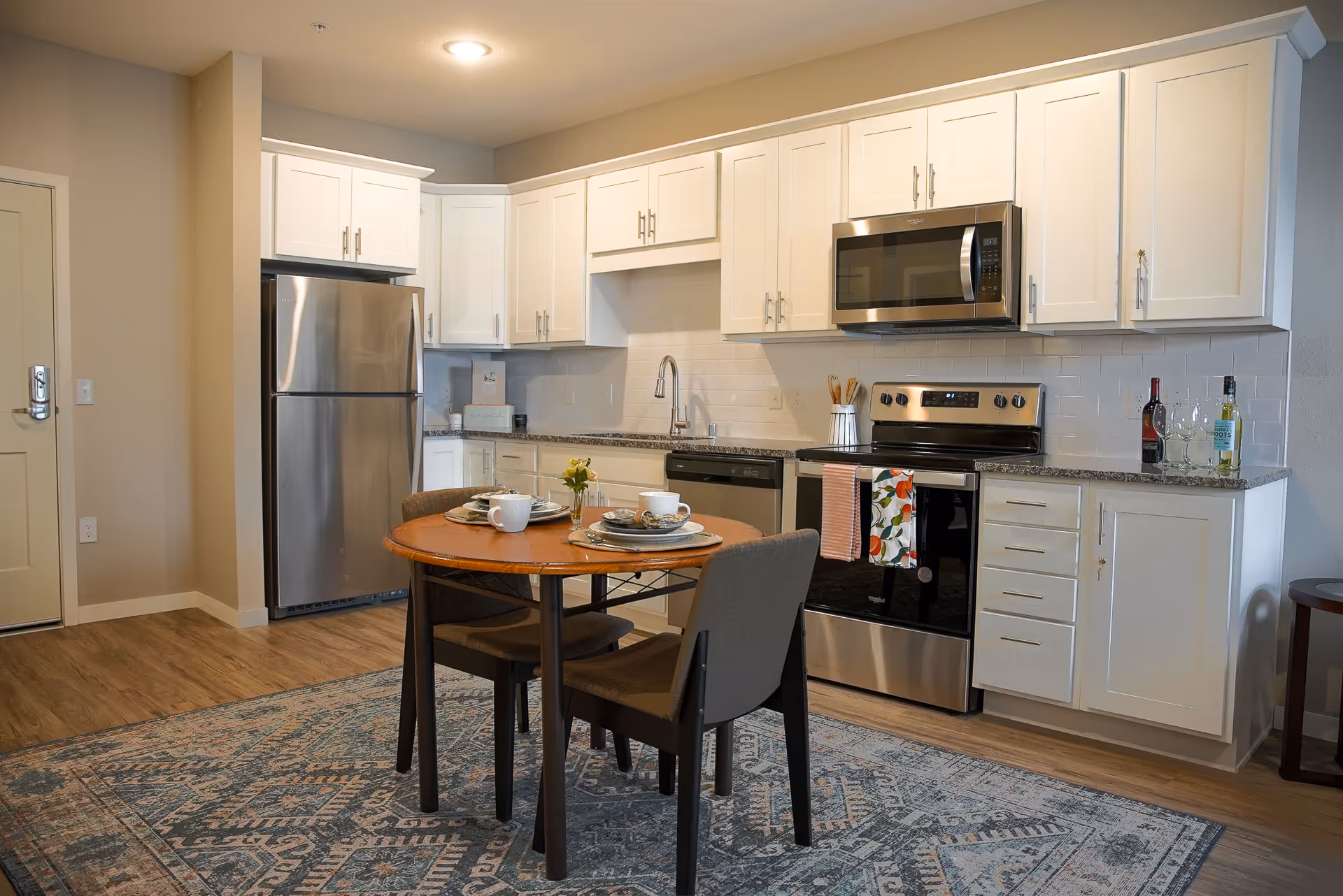 A modern kitchen with white cabinets, stainless steel refrigerator, oven, microwave, and dishwasher. A small round wooden dining table with two chairs is set with plates, cups, and a small flower vase. The floor has a patterned rug and the walls are painted beige.