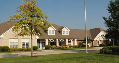 Exterior view of a single-story senior living facility building with a well-maintained lawn, a tree in the foreground, and patriotic red, white, and blue bunting hanging from the porch. The sky is clear and blue.