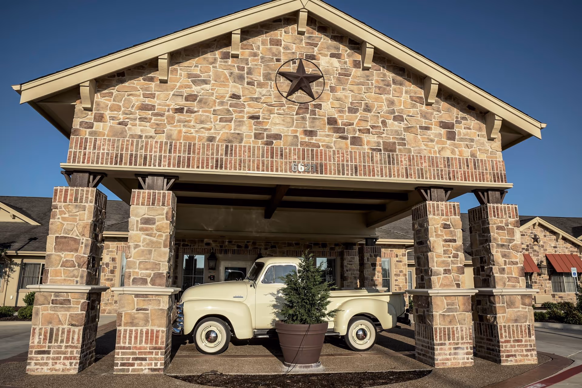 Stone entrance structure with four large stone pillars supporting a roof with a star emblem. A vintage cream-colored pickup truck is parked under the structure next to a large potted plant. The building exterior is made of stone and brick with a clear blue sky in the background.
