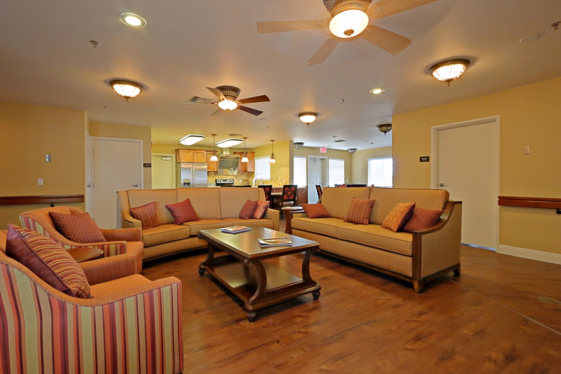 A spacious living room area with two beige sofas and two striped armchairs arranged around a wooden coffee table. The room has wooden flooring, ceiling fans with lights, and warm yellow walls. In the background, there is an open kitchen with wooden cabinets, stainless steel appliances, and a dining area with tables and chairs.
