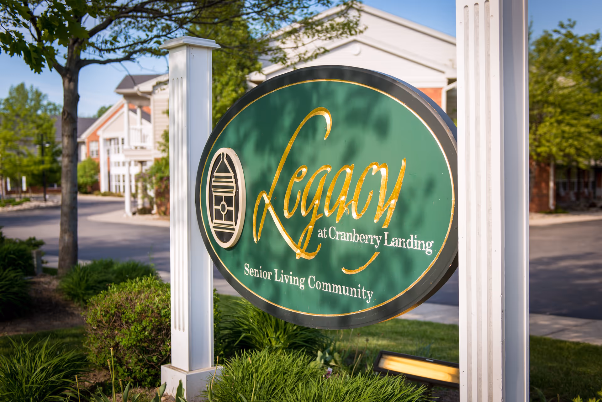 A green oval sign with gold lettering that reads 'Legacy at Cranberry Landing Senior Living Community' mounted on white posts, with a residential building and trees in the background under a clear blue sky.