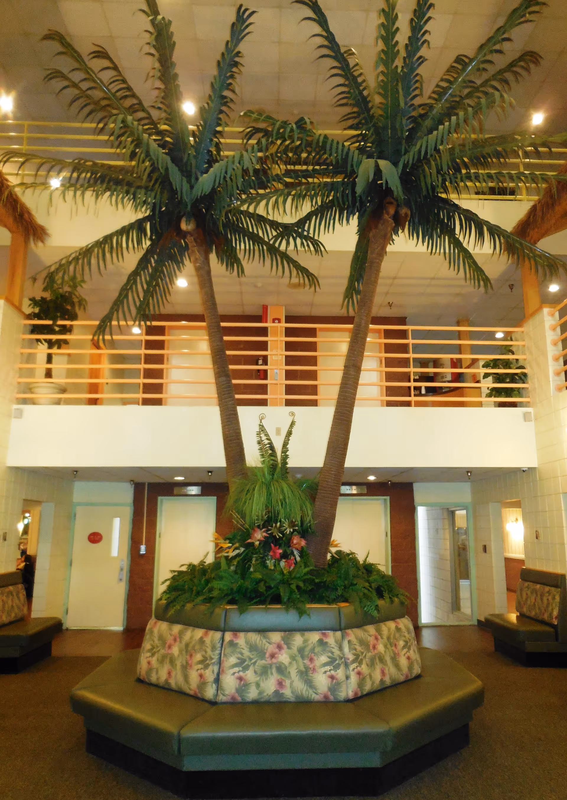 Indoor seating area with a large planter in the center containing two tall artificial palm trees and various green plants. The seating around the planter is upholstered with a floral pattern. The area has a high ceiling with a balcony railing above and doors leading to other rooms.