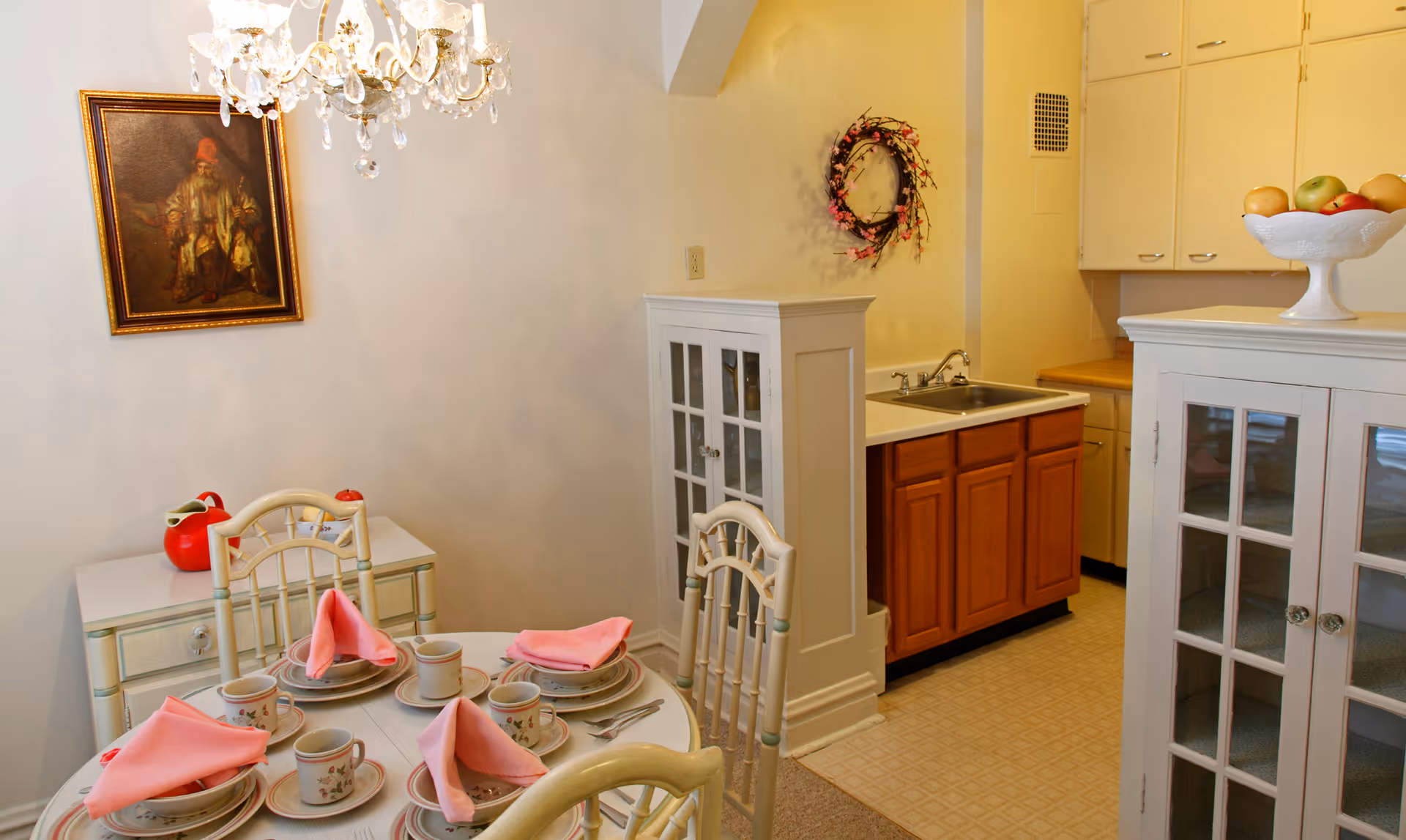 A cozy dining area with a round table set for four with pink napkins, cups, and plates. The room features a chandelier, a framed painting on the wall, and a small kitchen area with wooden cabinets and a sink. There are white cabinets with glass doors and a decorative fruit bowl on top.