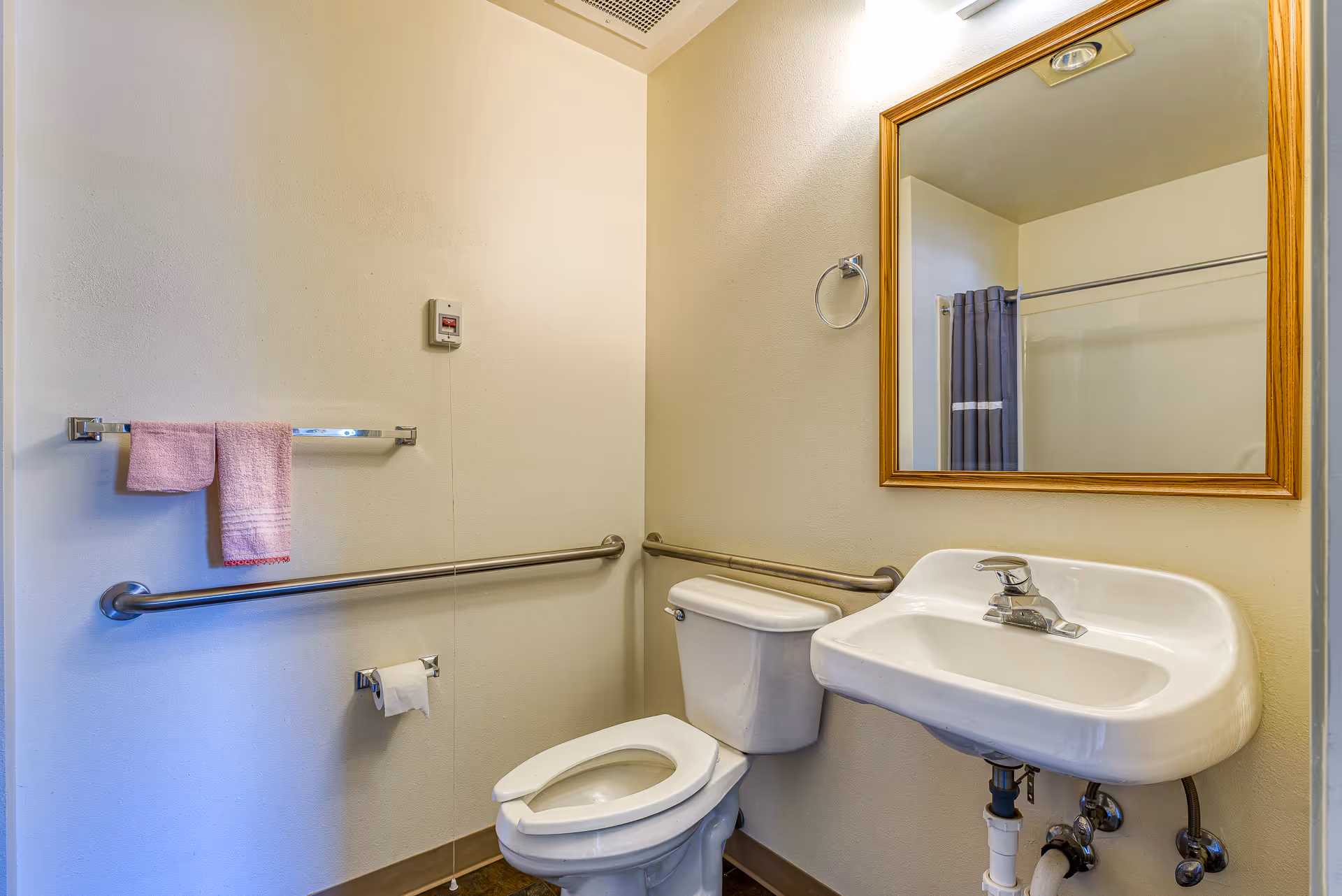 A small bathroom with a white toilet and a white wall-mounted sink. There are metal grab bars on the walls near the toilet and sink. A wooden framed mirror hangs above the sink. A towel rack with two pink towels and a toilet paper holder with a roll of toilet paper are mounted on the wall. A shower curtain is partially visible in the mirror reflection.
