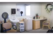 Interior view of a salon or grooming area with two salon chairs, a hair dryer, a sink, a large mirror, a small storage drawer unit, a countertop with various items, and a decorative wreath on the wall.
