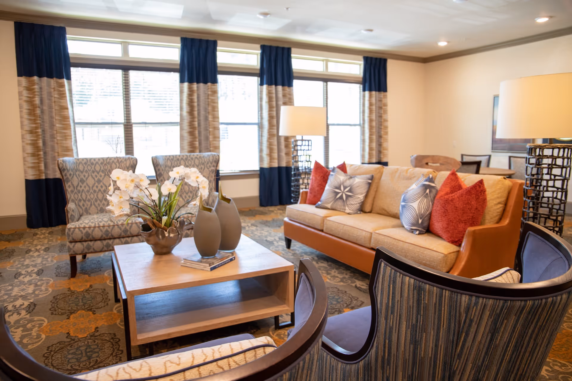 A bright and cozy living room with a beige sofa adorned with red and patterned throw pillows, two patterned armchairs, and two striped chairs around a wooden coffee table decorated with white orchids and two modern vases. Large windows with blue and beige curtains allow natural light to fill the room, and a floor lamp stands beside the sofa. In the background, there is a dining table with chairs and a framed picture on the wall.