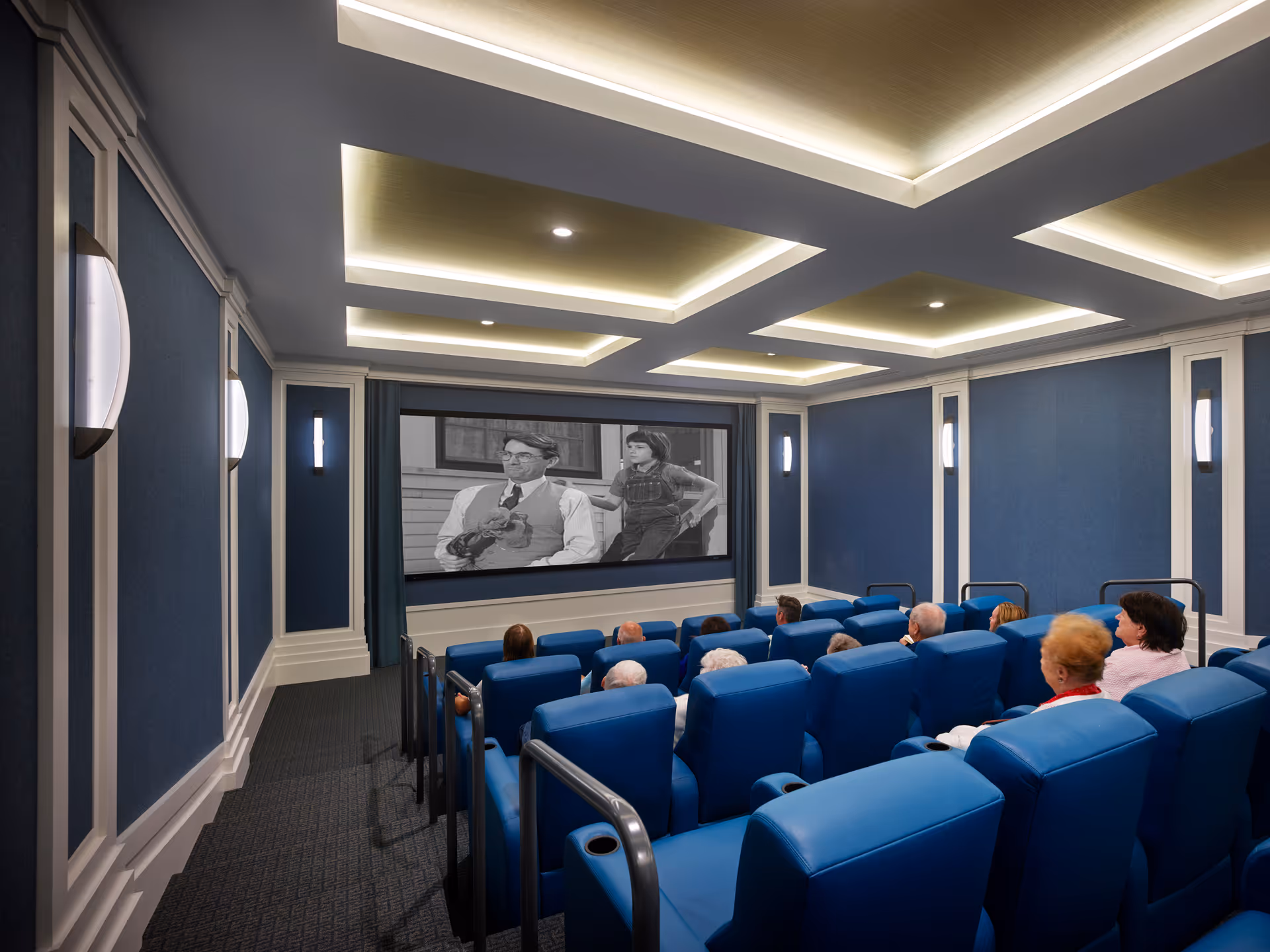 Small theater-style screening room with rows of blue recliner seats and seniors watching a black-and-white movie on a front screen.