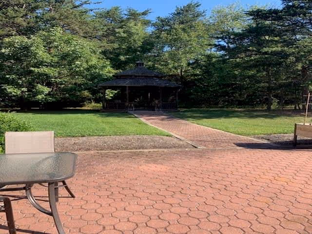 Outdoor patio area with a glass table and chairs in the foreground, a brick pathway leading to a gazebo surrounded by green grass and trees under a clear blue sky.